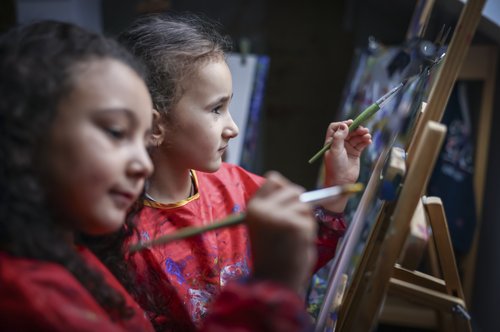 Aktivität in der Kita: Zwei Mädchen im Kindergartenalter sitzen im Malatelier vor ihren Staffeleien und malen mit Pinsel und viel Farbe kreative Bilder.