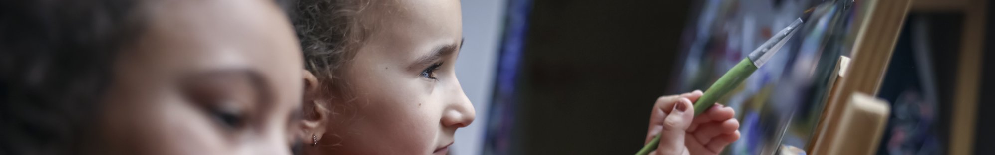 Aktivität in der Kita: Zwei Mädchen im Kindergartenalter sitzen im Malatelier vor ihren Staffeleien und malen mit Pinsel und viel Farbe kreative Bilder.