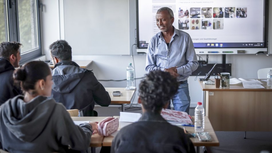 Unterrichtssituation im Hauptschul- und Berufsvorbereitungslehrgang: Die Lehrkraft spricht mit den jugendlichen Schülerinnen und Schülern, die mit dem Rücken zur Kamera sitzen, im Hintergrund das Smartboard mit Unterrichtsmaterial.