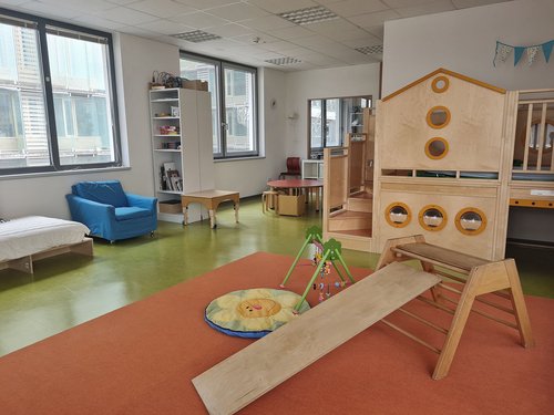 Großes, helles, betreutes Kinderzimmer Bekidz in der Frankfurt University of Applied Sciences, mit grünem Fußboden, zwei Fenstern, blauem Sessel, einem weißen Regalschrank mit Büchern und Spielen, einer Holzspielebene mit Treppenstufen und einer Krabbeldecke mit Spielbogen.