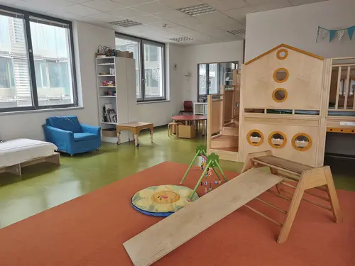 Großes, helles, betreutes Kinderzimmer Bekidz in der Frankfurt University of Applied Sciences, mit grünem Fußboden, zwei Fenstern, blauem Sessel, einem weißen Regalschrank mit Büchern und Spielen, einer Holzspielebene mit Treppenstufen und einer Krabbeldecke mit Spielbogen.