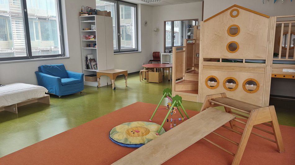 Großes, helles, betreutes Kinderzimmer Bekidz in der Frankfurt University of Applied Sciences, mit grünem Fußboden, zwei Fenstern, blauem Sessel, einem weißen Regalschrank mit Büchern und Spielen, einer Holzspielebene mit Treppenstufen und einer Krabbeldecke mit Spielbogen.