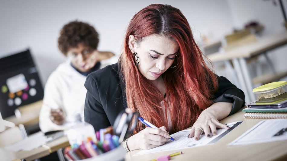 Im Vorkurs für den Hauptschul- und Berufsvorbereitungslehrgang schreibt eine junge rothaarige Frau konzentriert in ihrem Block, während ihr Klassenkamerad im Hintergrund (verschwommen) in Gedanken versunken in die Kamera blickt.